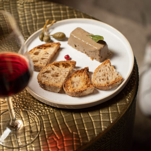 Gourmet appetizer with pâté, toasted bread, and capers served at Oratio Wine Bar & Library, paired with a glass of red wine.