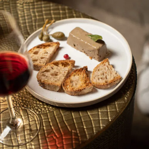 Gourmet appetizer with pâté, toasted bread, and capers served at Oratio Wine Bar & Library, paired with a glass of red wine.